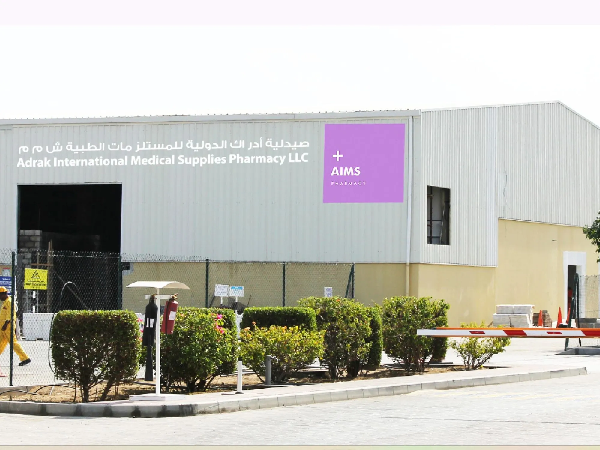 Exterior view of Adrak International Medical Supplies Pharmacy warehouse, with a sign saying "AIMS Pharmacy." The building is surrounded by a fence and parking barriers.